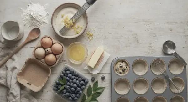 Overhead flat-lay view of lemon blueberry muffin ingredients including flour, lemon zest sugar, eggs, butter, and fresh blueberries