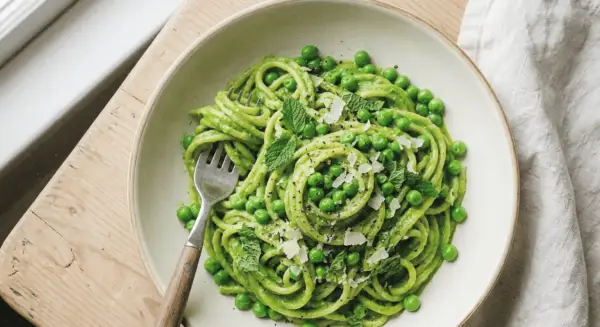 Overhead shot of creamy green pasta in a shallow bowl, flecked with peas and fresh herbs, twirled with a fork.