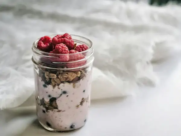 Clear glass breakfast jar of creamy overnight oats soaked in milk and yoghurt, topped with fresh raspberries in soft morning daylight