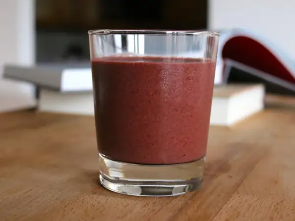 A cold glass of deep purple blackberry banana smoothie with visible tiny berry flecks on a wooden table