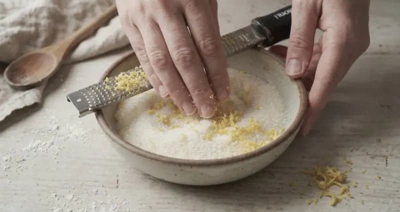 Close-up shot of hands rubbing fresh lemon zest directly into white granulated sugar in a ceramic bowl