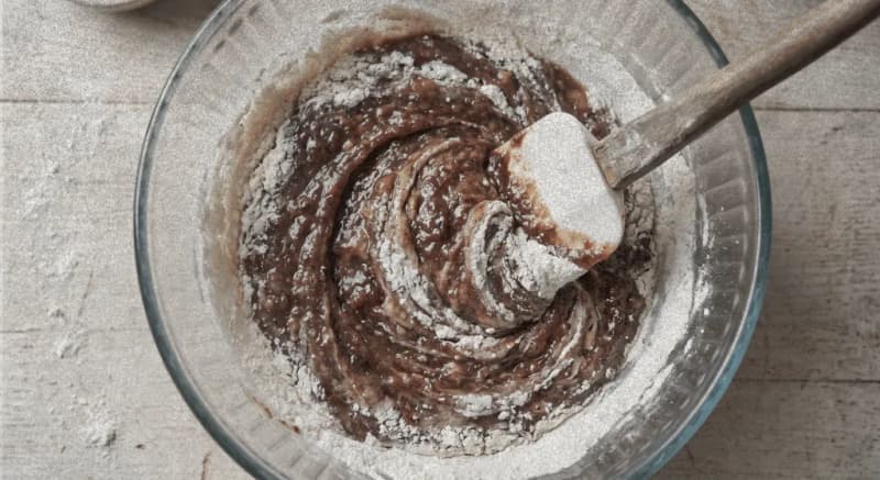 Action shot of a spatula folding dry flour into moist banana batter in a glass bowl, no berries visible