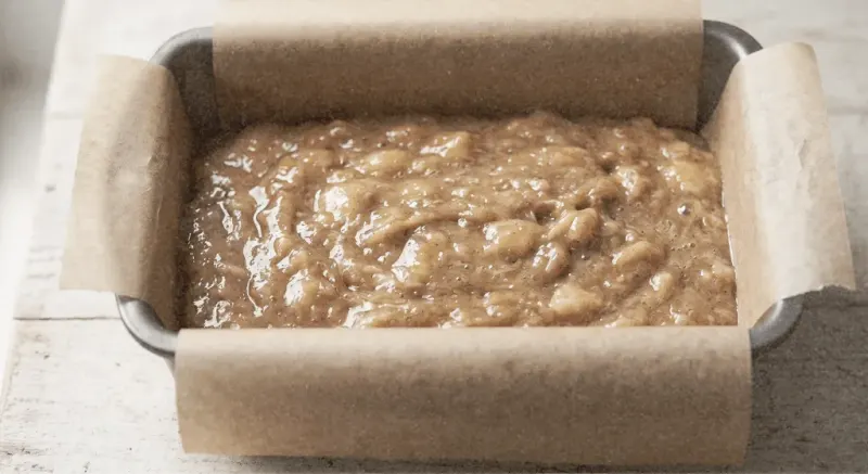 Angled close-up of thick banana loaf batter poured into a parchment-lined metal loaf pan ready for the oven, plain top